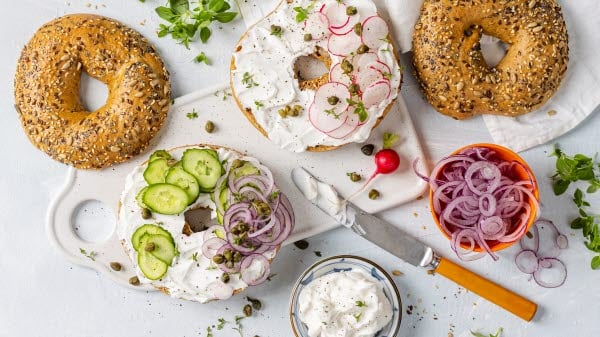 Fresh bagels spread with cream cheese made from plant based dairy, garnished with cucumber slices and onion rings, presented on a white plate.
